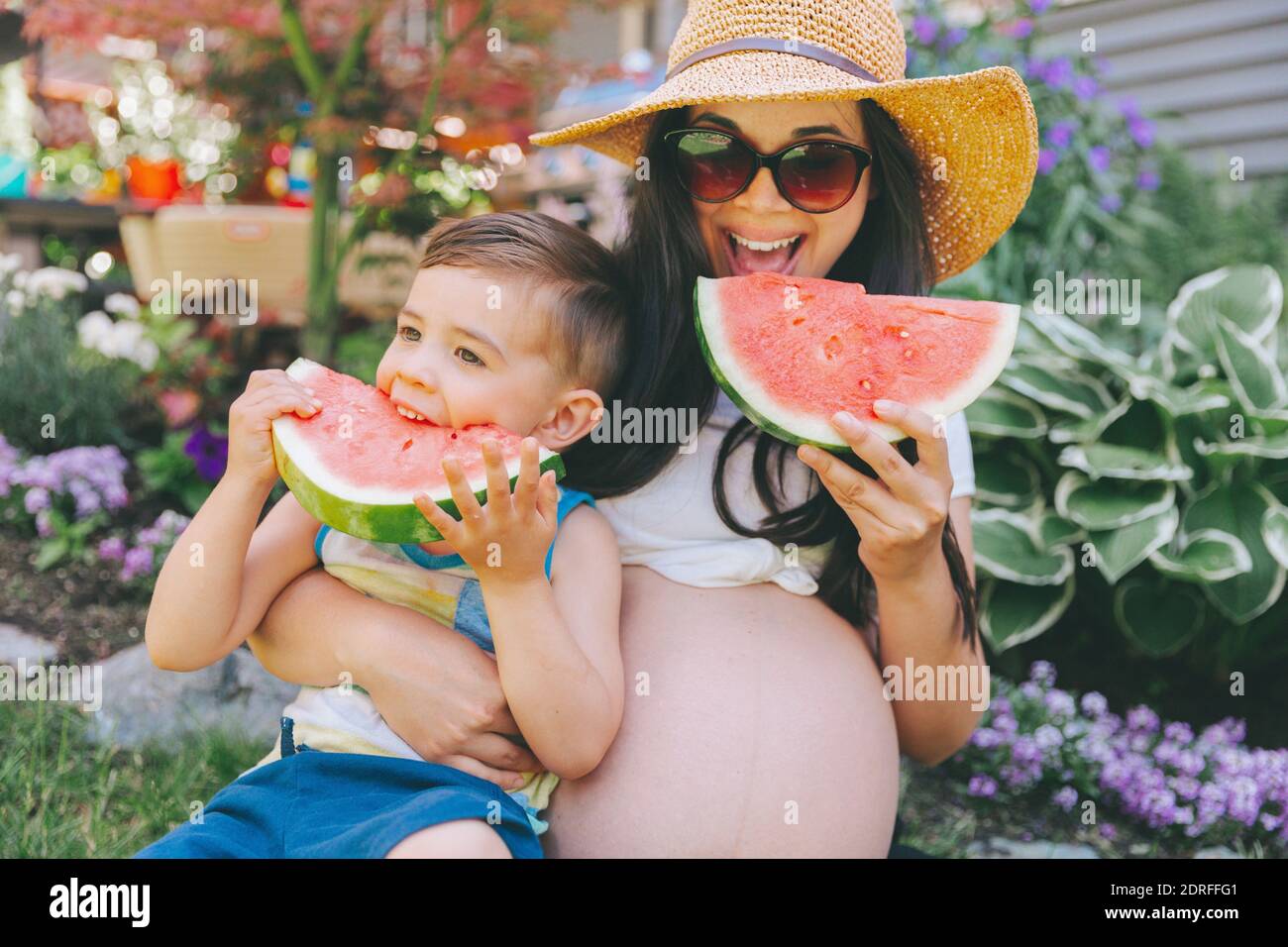 Pregnant Mother And Son Eating Watermelons While Sitting Against Plants