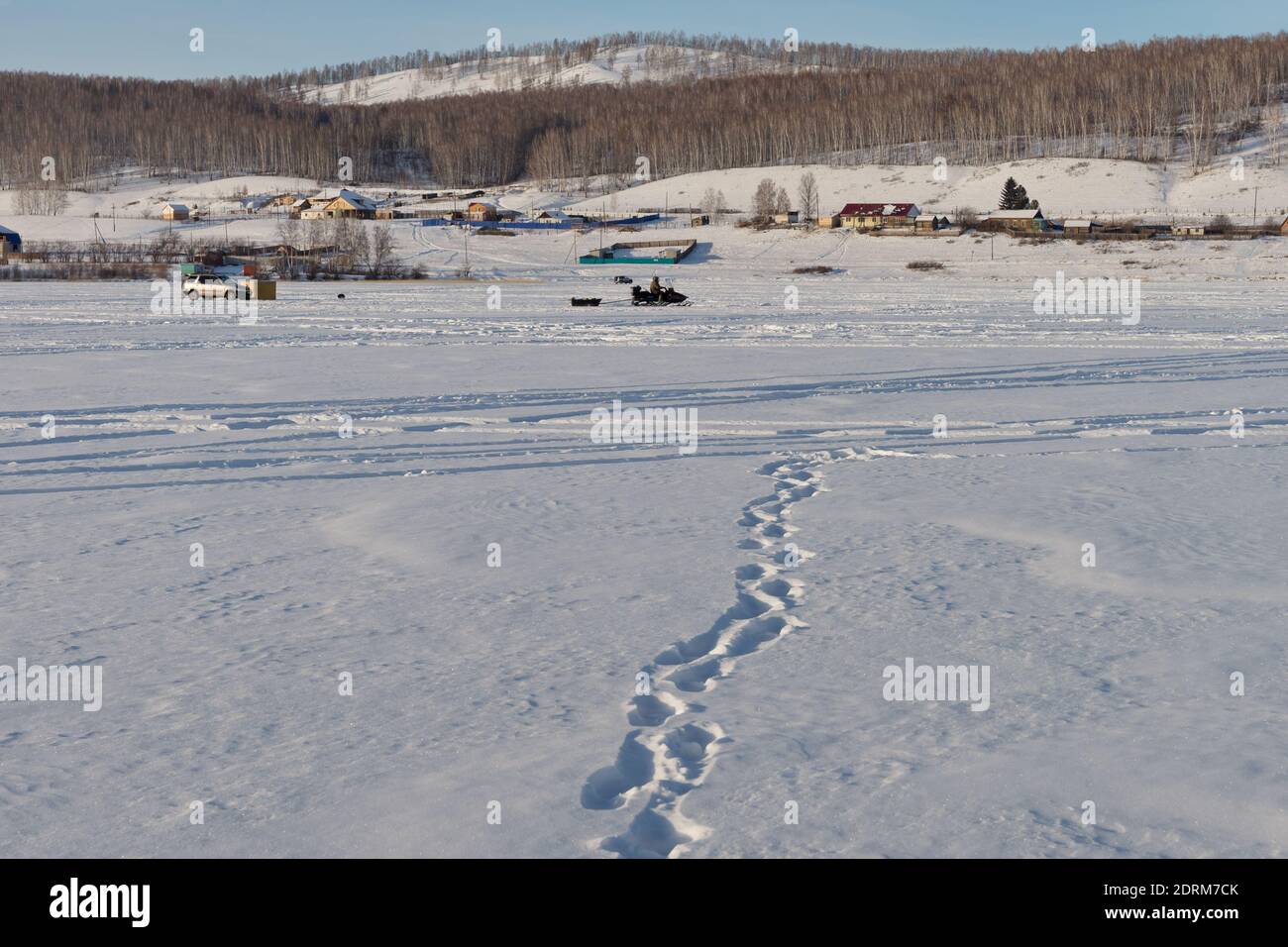 Human footprints lead to the rural shore along the frozen snow-covered surface of a large lake. Stock Photo