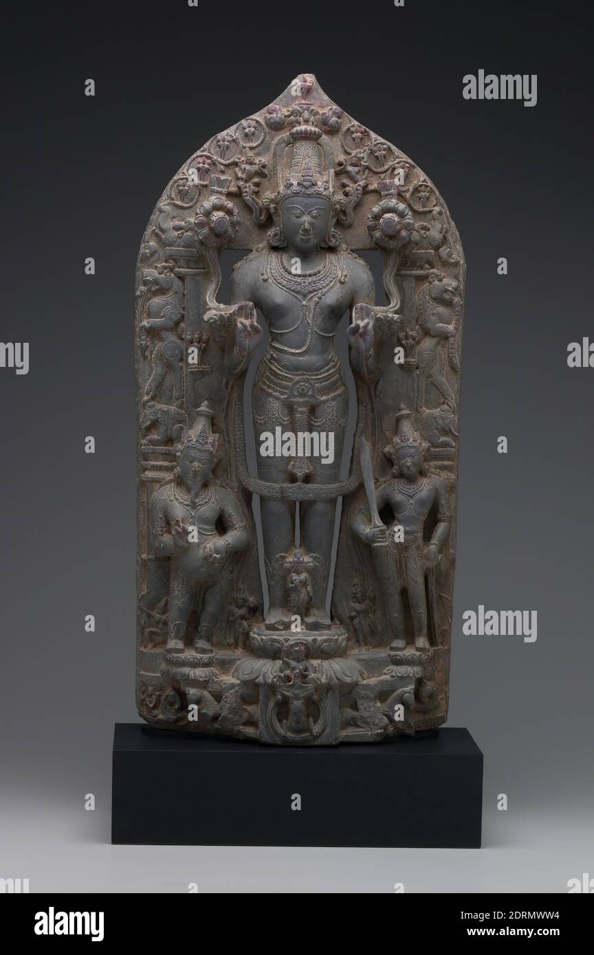 Sun God Surya with Adityas, Attendants, and Wives, late 11th century, Schist, 74.9 × 35.6 × 14 cm (29 1/2 × 14 × 5 1/2 in.), Surya is featured in both Buddhist and Hindu traditions and is one of the twelve Adityas, or children of the goddess of the heavens. Identified by the horse-drawn chariot at the bottom of this sculpture, he holds lotuses in his two hands and is surrounded by smaller representations of his eleven brothers. Pingala, the potbellied and bearded recorder, stands to his right, and Danda, the measurer, stands to his left holding a sword. Stock Photo