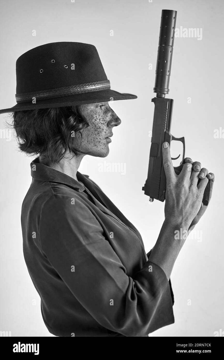 female with weapon against white background, isolated. detective redhead woman in black hat looking away, applying the weapon's barrel up Stock Photo