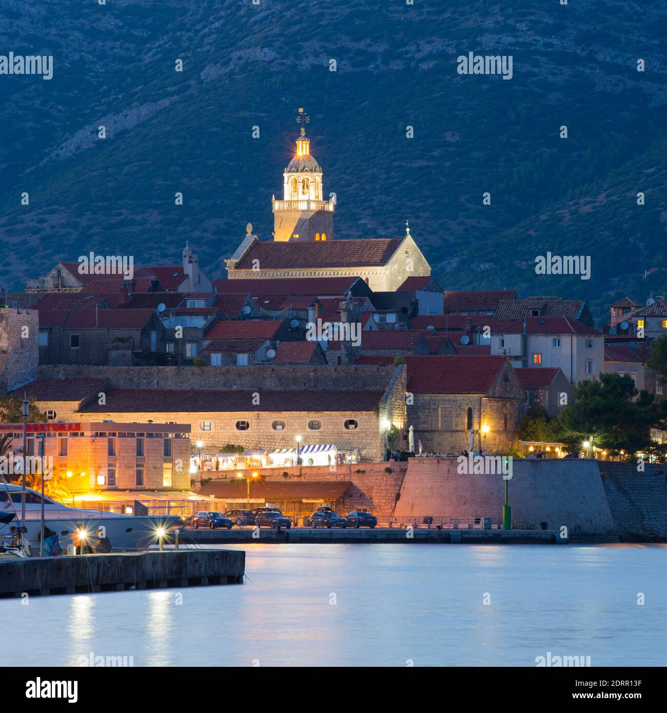 Korčula Town, Korčula, Dubrovnik-Neretva, Croatia. View across marina to the illuminated Old Town, dusk, St Mark's Cathedral prominent. Stock Photo