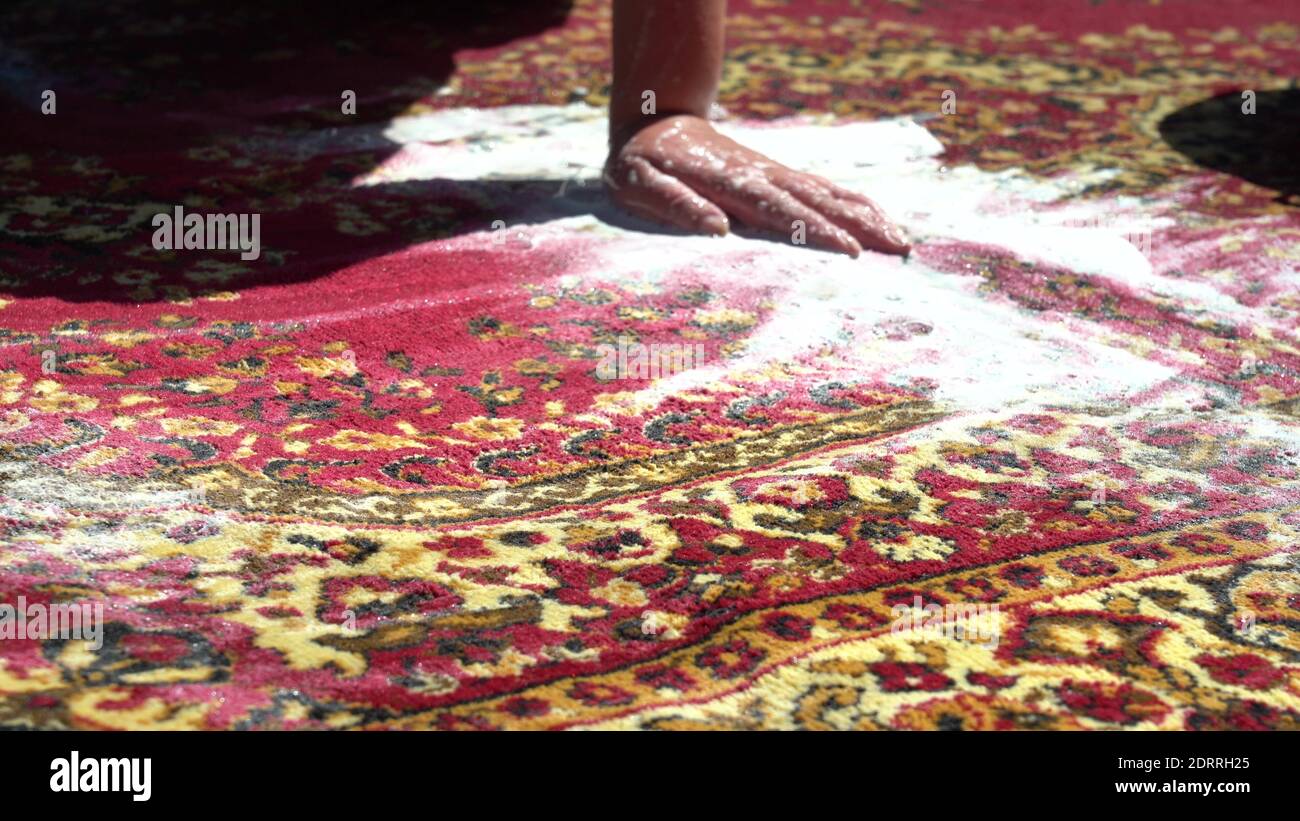 A woman is brushing a dirty carpet in the street, in the courtyard of the house close up. Rinse with clean water. A lot of foam from the powder Stock Photo