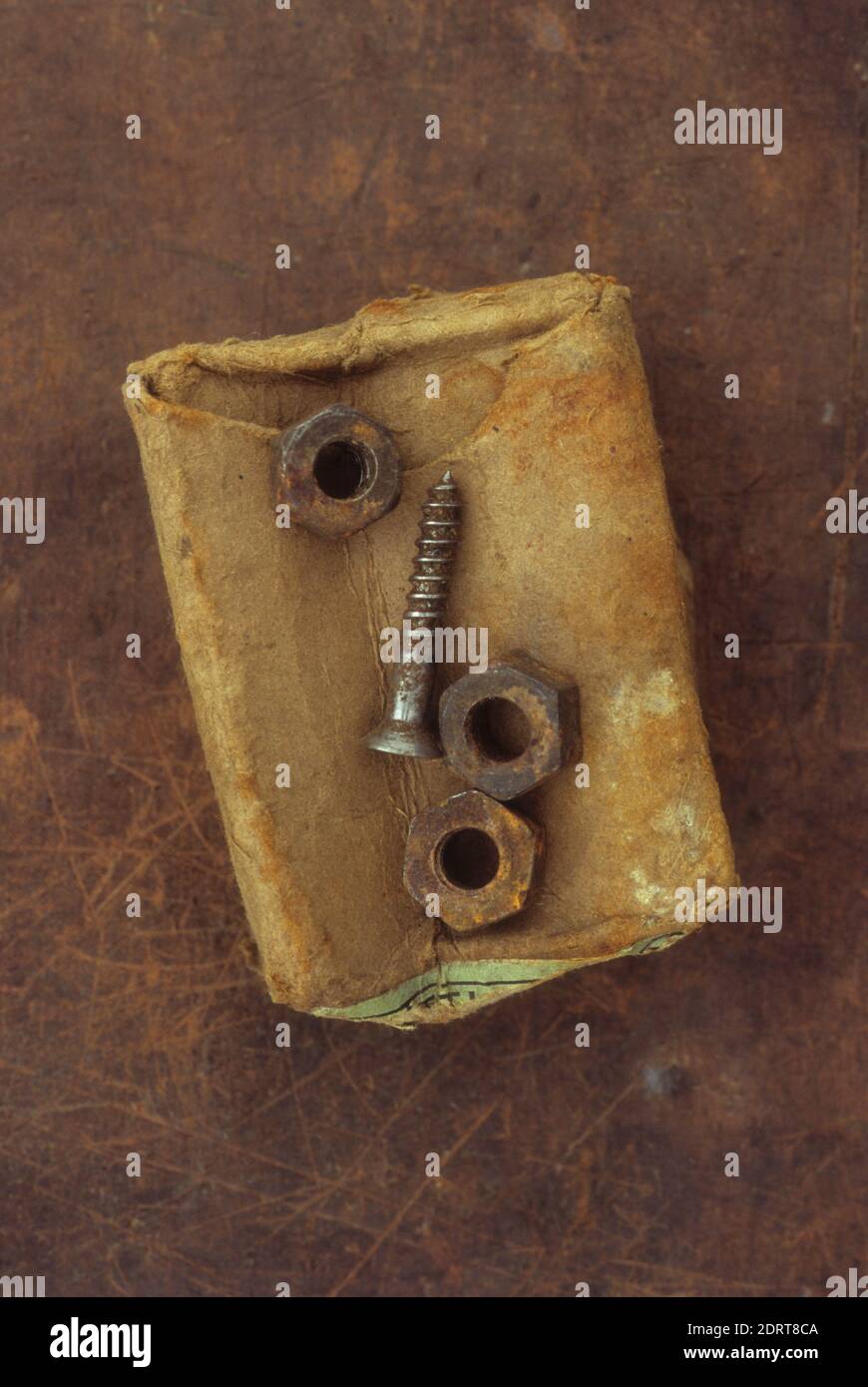 Old crumpled rust-stained cardboard box with three rusty metal nuts and used screw lying on top Stock Photo
