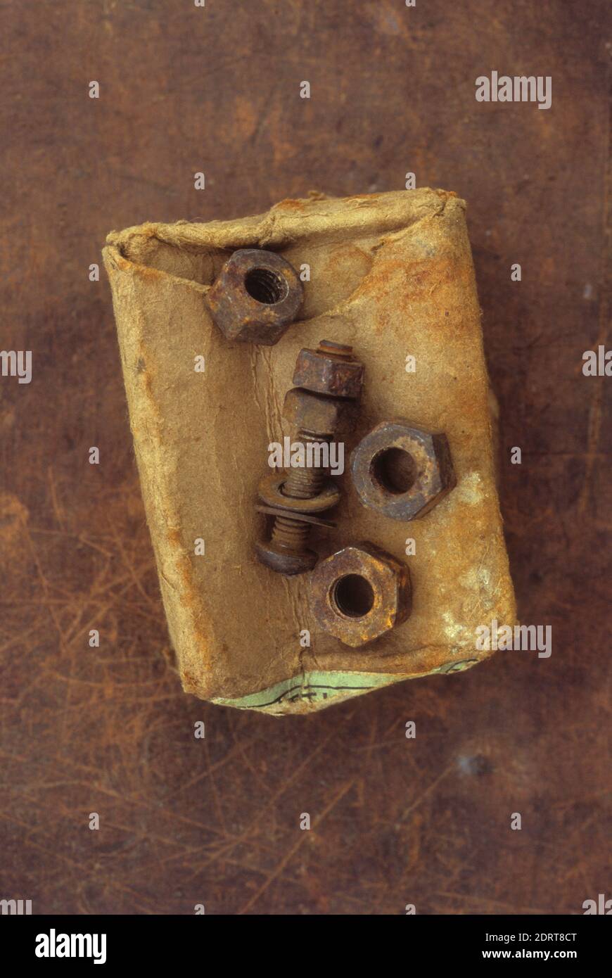 Old crumpled rust-stained cardboard box with three rusty metal nuts and used screw lying on top Stock Photo