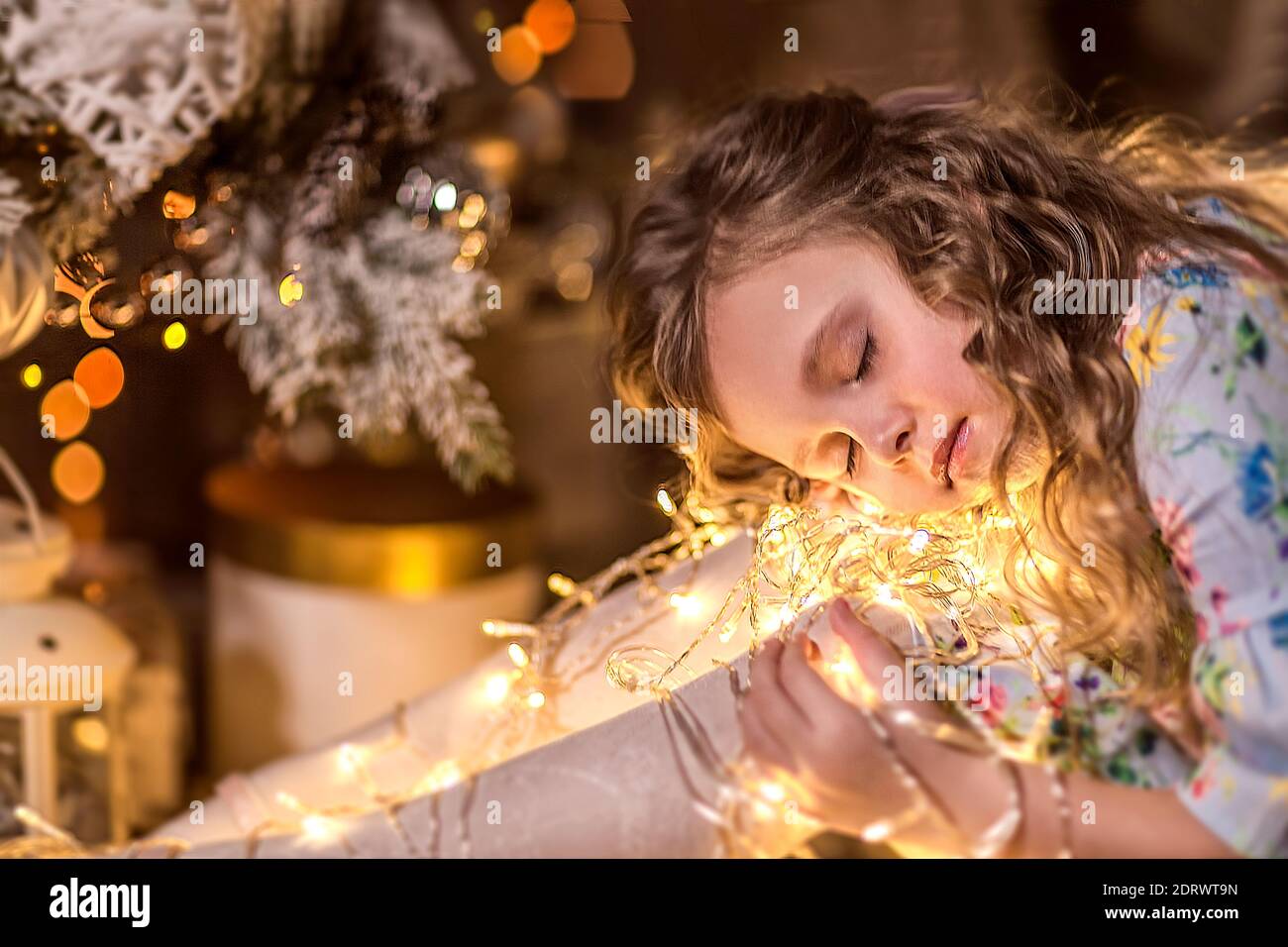A beautiful girl with curly long hair is sitting near a Christmas tree. Holding a garland of Christmas lights. The concept of winter recreation at hom Stock Photo