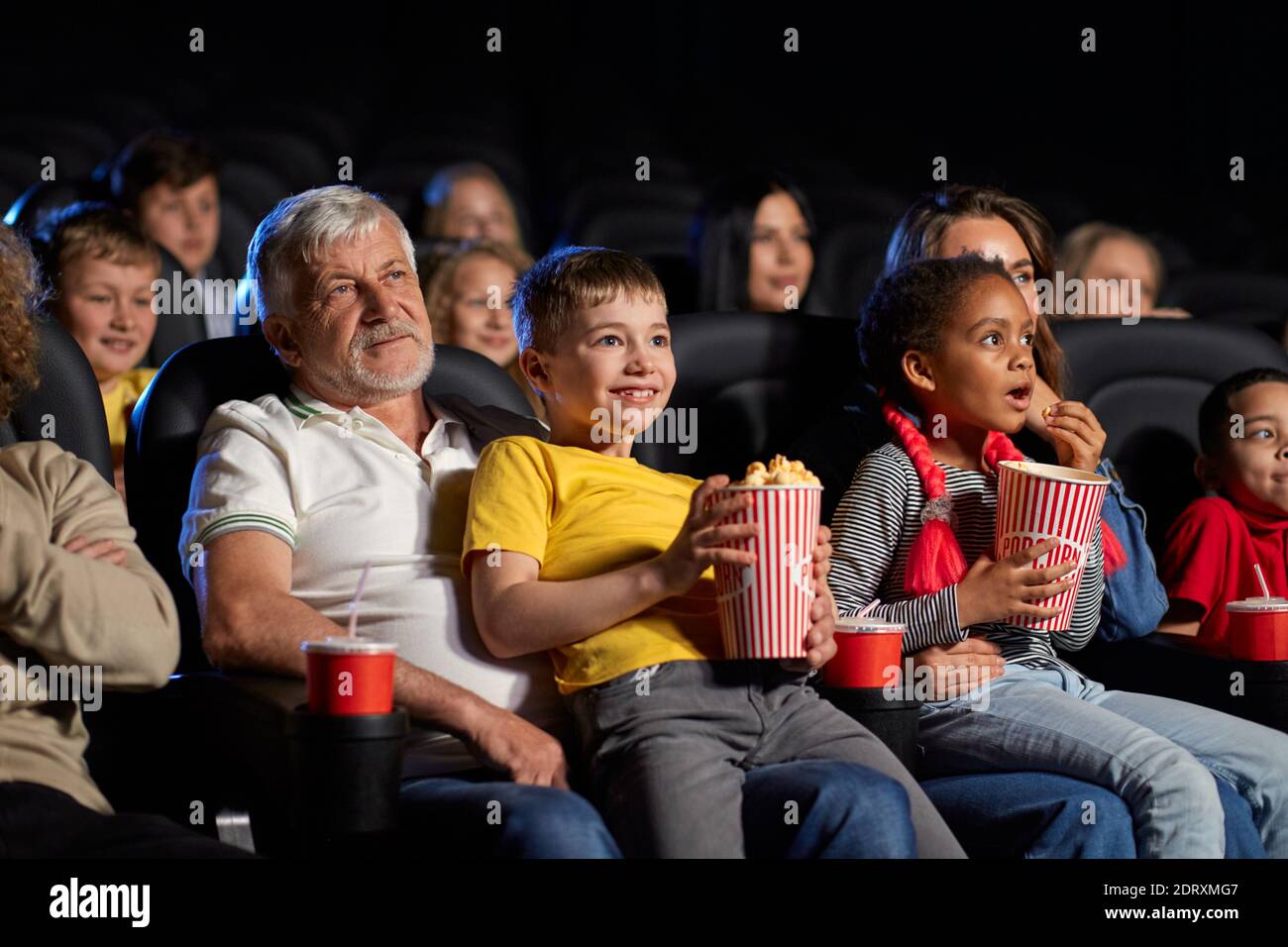 Little african girl with caucasian woman and caucasian boy with senior grandfather. Side view of multiracial kids holding popcorn and sitting on knees Stock Photo