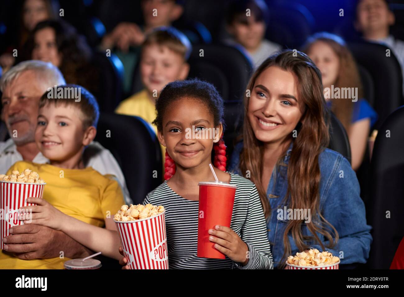 Mother enjoying time with half african little daughter, kid sitting on knees, holding drink and popcorn, looking at camera. Happy multiracial family w Stock Photo