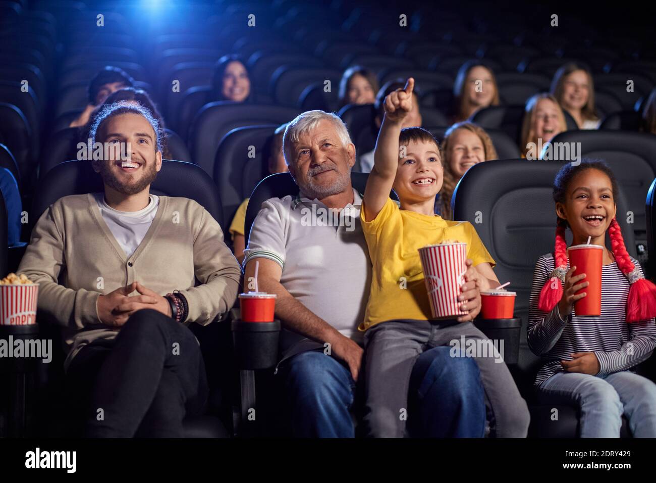 Bearded grandfather with grandson sitting on knees, child holding popcorn and pointing at screen, young multiracial audience laughing. Happy family en Stock Photo