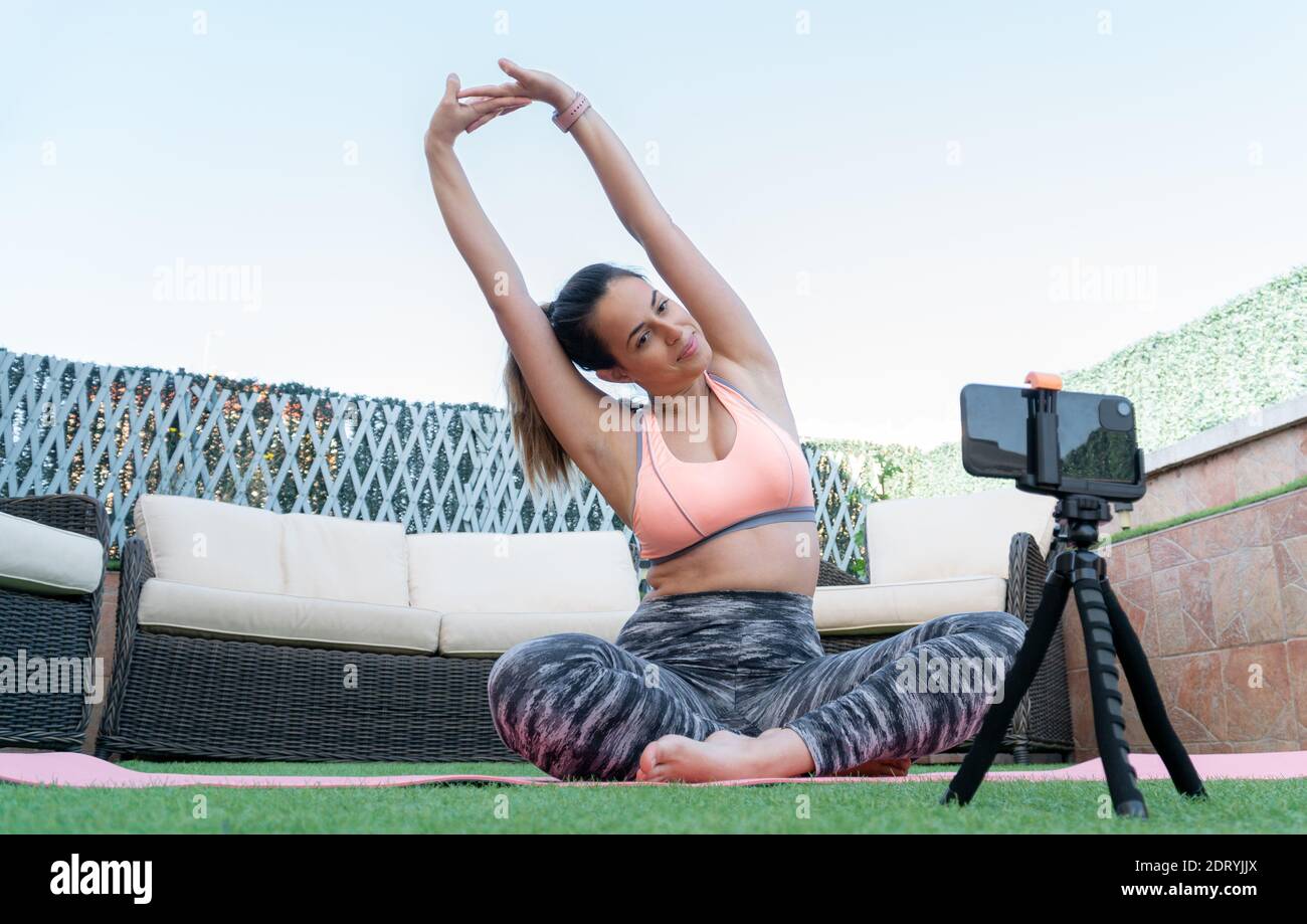 Active young woman, vlogger does sports, records her workout training from  home on smartphone camera, posing for selfie inside her house, sits on  rubber yoga mat in blue leggings and sportsbra 35338950