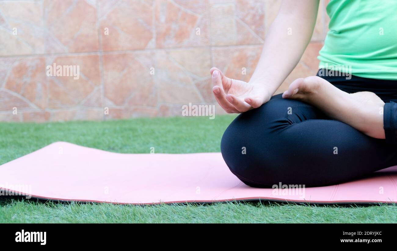 A woman doing yoga in the garden of her house. We can see a close-up of her right leg and arm while the woman performs meditation exercises on a pink Stock Photo