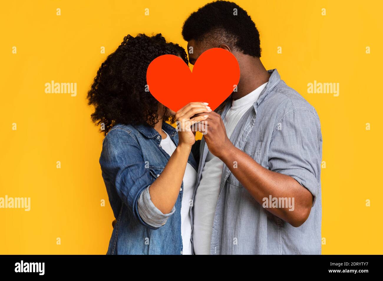 Secret Love. Romantic black couple hiding and kissing behind red paper heart Stock Photo