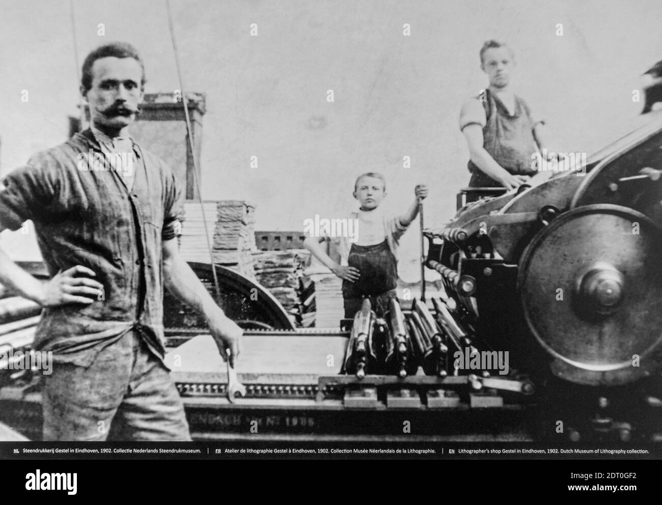 1902 black and white archival photo showing pressman and children working on lithographic printing press in pressroom / print shop Stock Photo