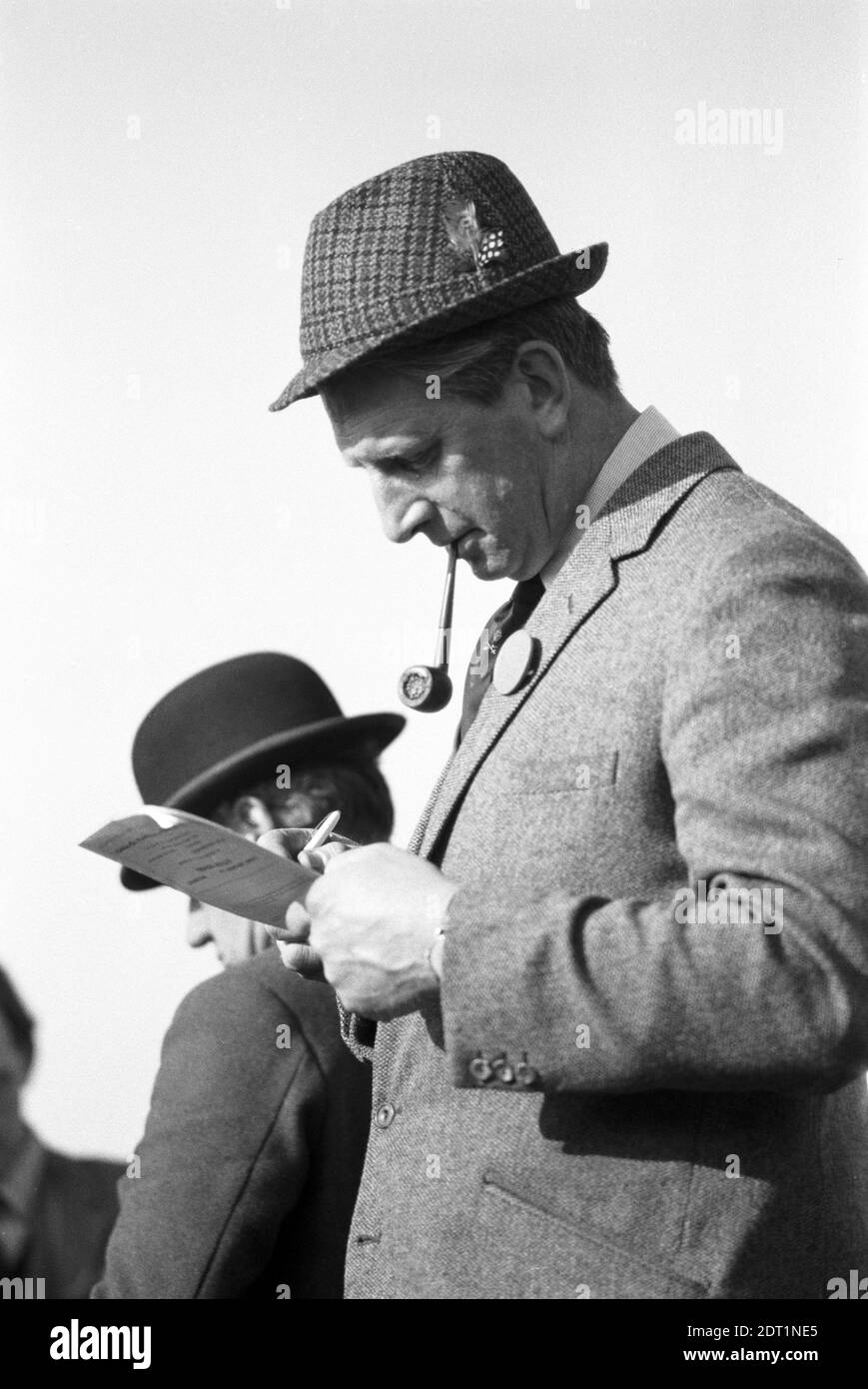 UK, England, Devonshire, Buckfastleigh, 1972. Point-to-Point races were held at  Dean Court on the Dean Marshes, close to the A38 between Plymouth and Exeter. A steward (with a badge) of the races studying the form on a racecard wearing a traditional wool tweed trilby hat with feathers in the band. The steward behind him is wearing a Bowler Hat. Stock Photo