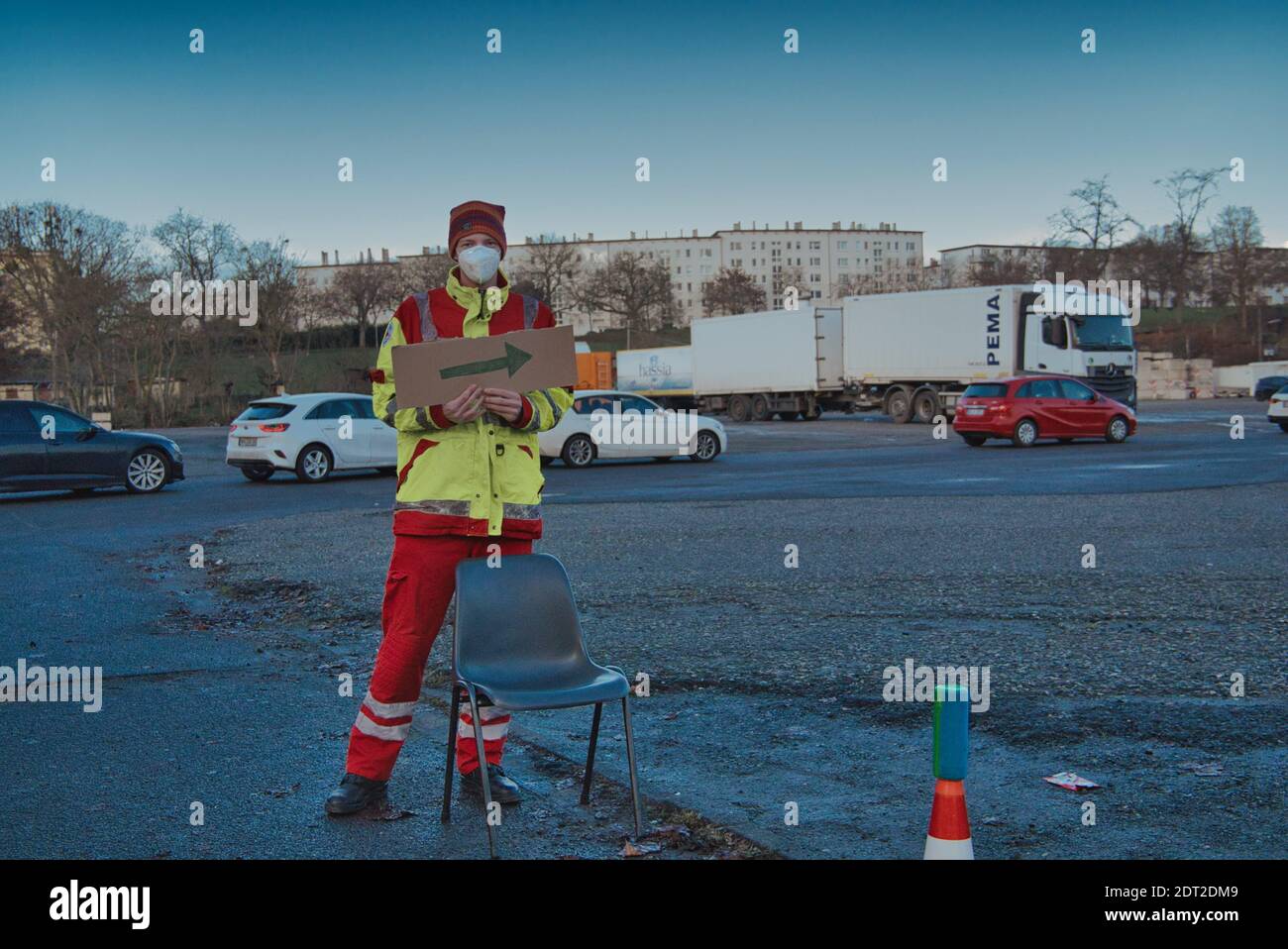 Frankfurt, Germany. December 2020. Young man wearing mask coveralls and safety vest holding cardboard direction arrow sign outdoors. Long shot. Stock Photo