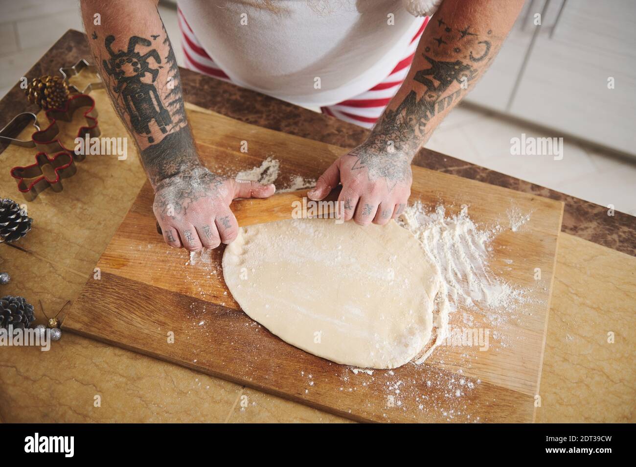 A man with a tattoo on his arms rolls out a flat dough on a wooden board Stock Photo