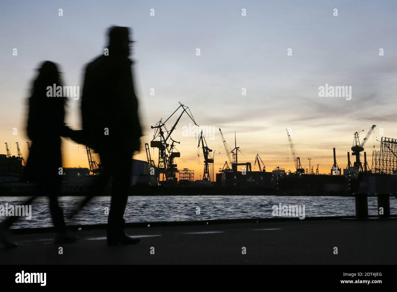 SpaziergŠnger sind bei Sonnenuntergang  an den LandungsbrŸcken unterwegs. Der abendliche Himmel ist hinter KrŠnen der Werft von Blohm + Voss im Hambur Stock Photo