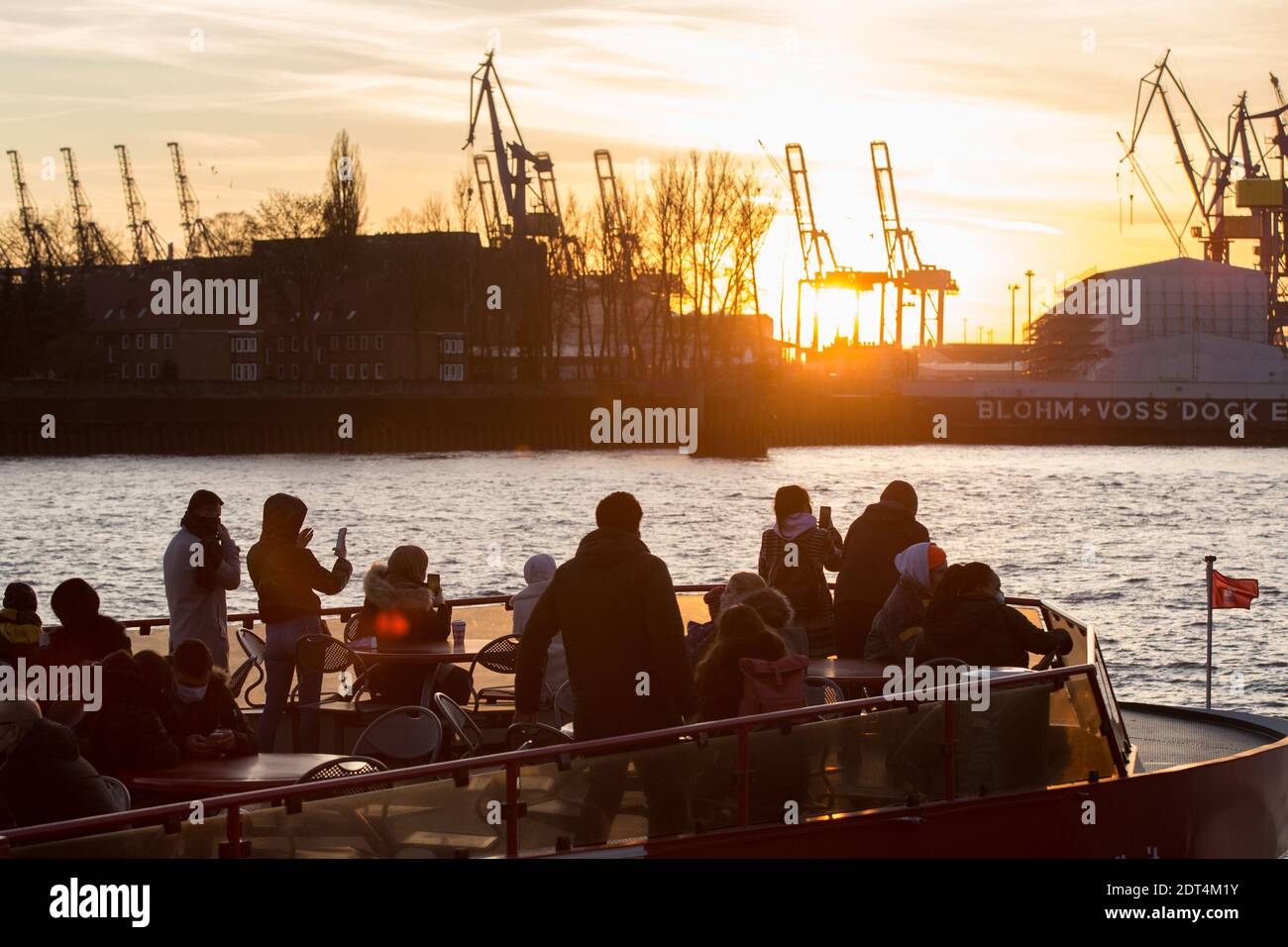 Menschen fahren bei Sonnenuntergang auf einer FŠhre auf der Elbe an den LandungsbrŸcken im Hafen von Hamburg. Auf der anderen Elbseite sind die KrŠne Stock Photo