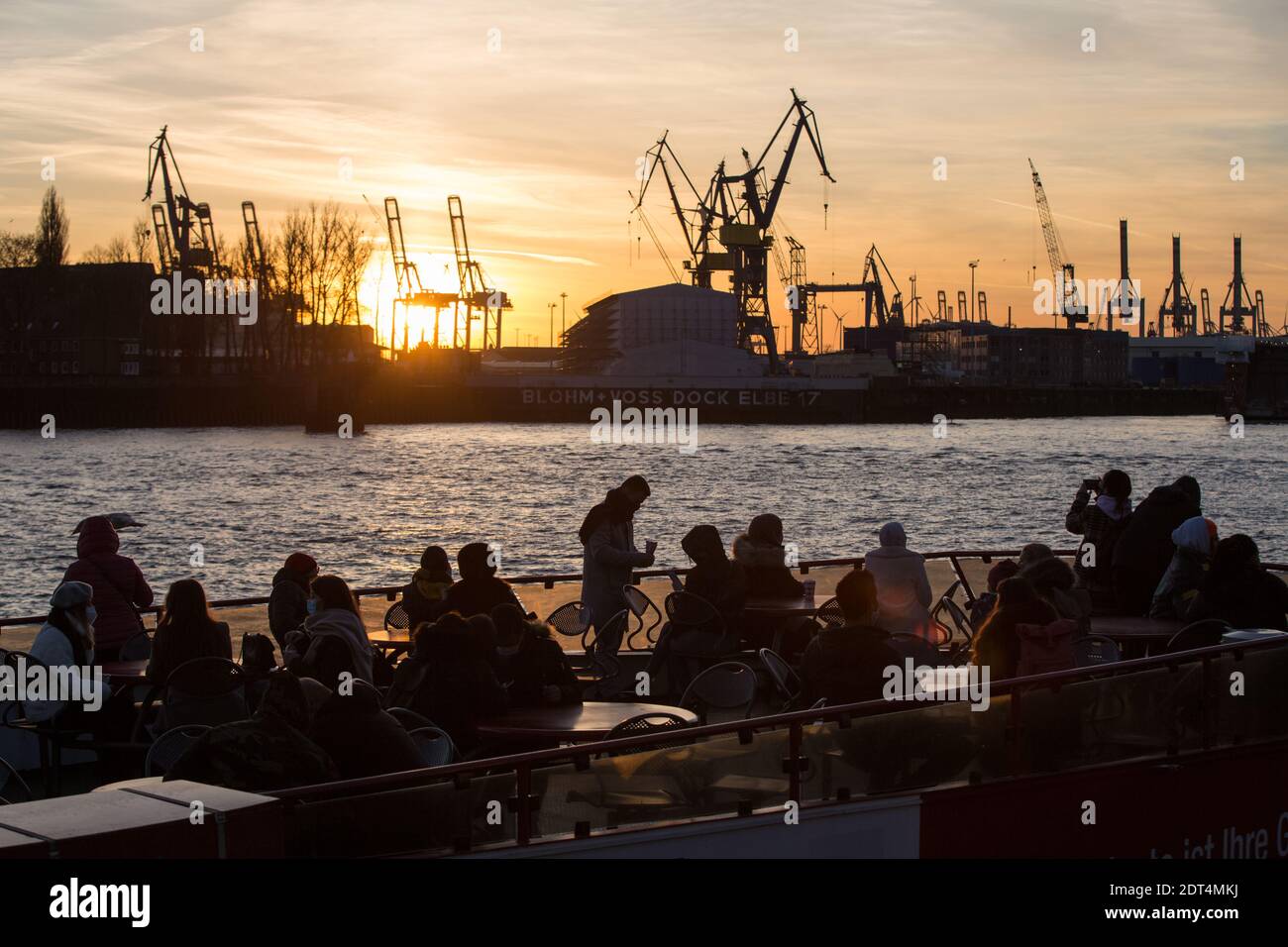 Menschen fahren bei Sonnenuntergang auf einer Fähre auf der Elbe an den Landungsbrücken im Hafen von Hamburg. Auf der anderen Elbseite sind die Kräne Stock Photo