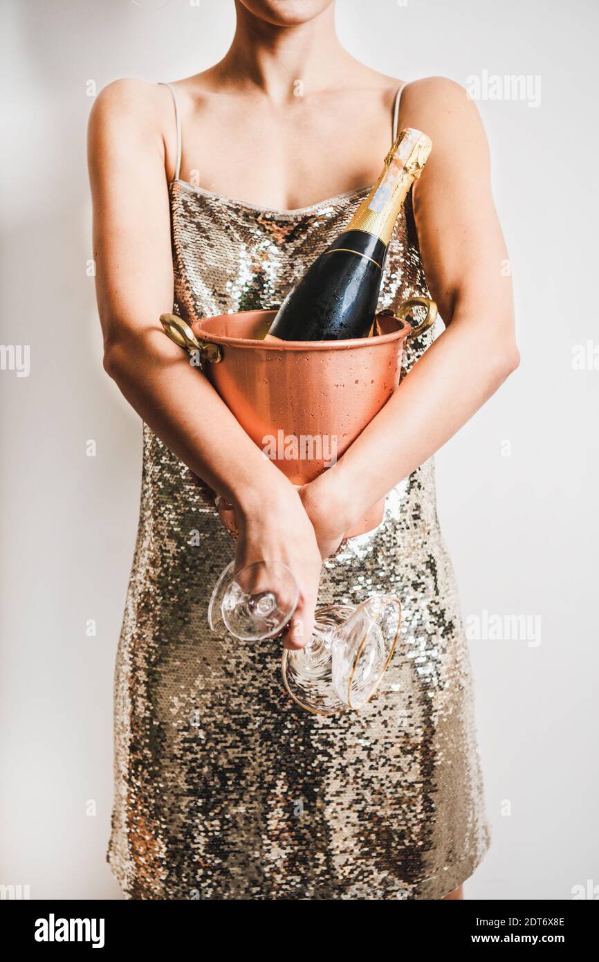 Young woman figure in festive glittering cocktail golden mini dress holding copper bucket with ice for champagne bottle and glasses over white wall background. New Year holiday party concept Stock Photo