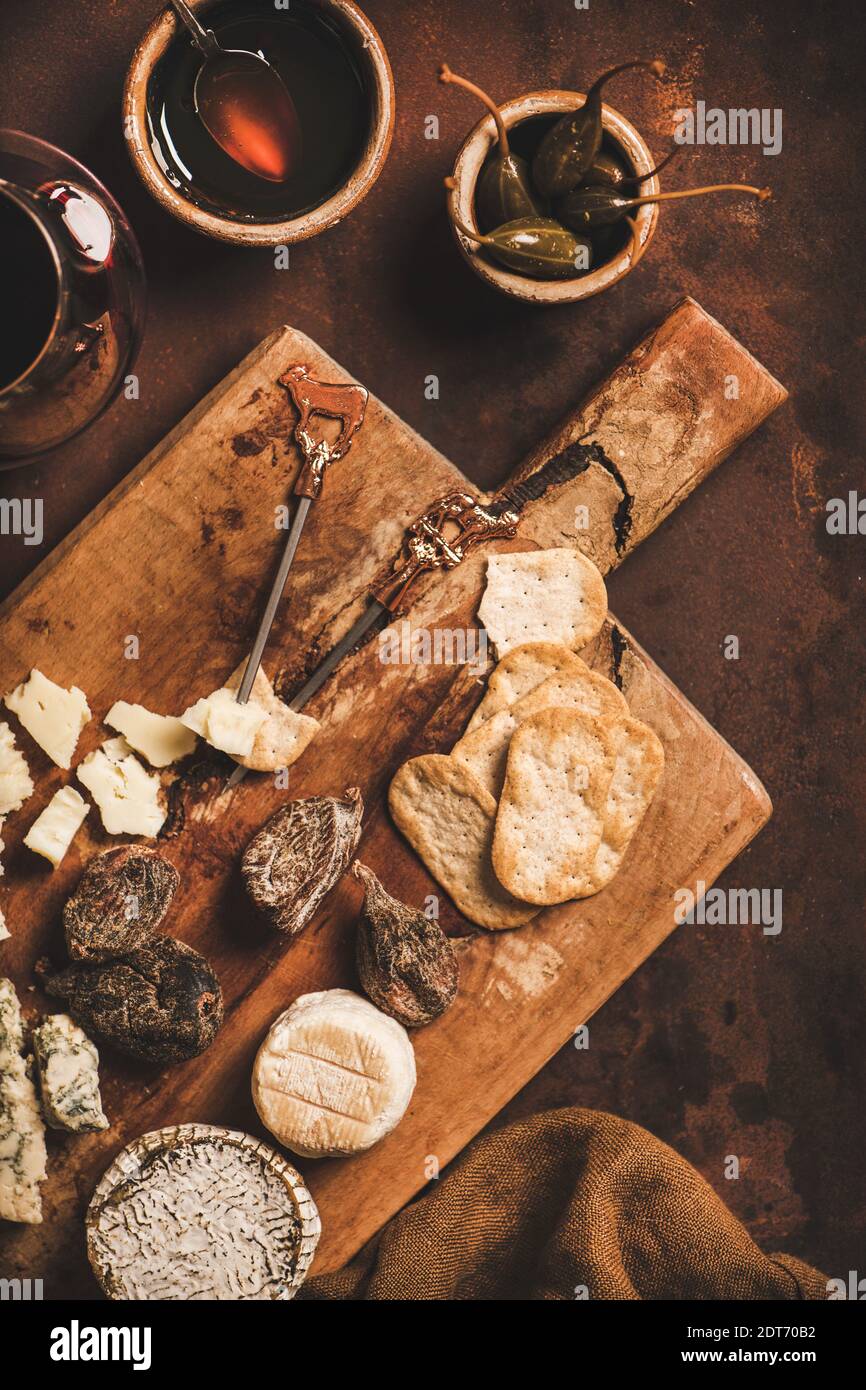 Assortment of snacks and appetizers for red wine concept. Flat-lay of various cheeses, crackers, smoked meat, fruit and glass of red wine on wooden rustic board over rusty brown background, top view Stock Photo