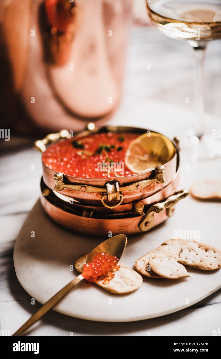 Red caviar in copper metal dishes with salty crackers and glass of champaigne on board over white marble table, selective focus. Winter holiday mood concept Stock Photo