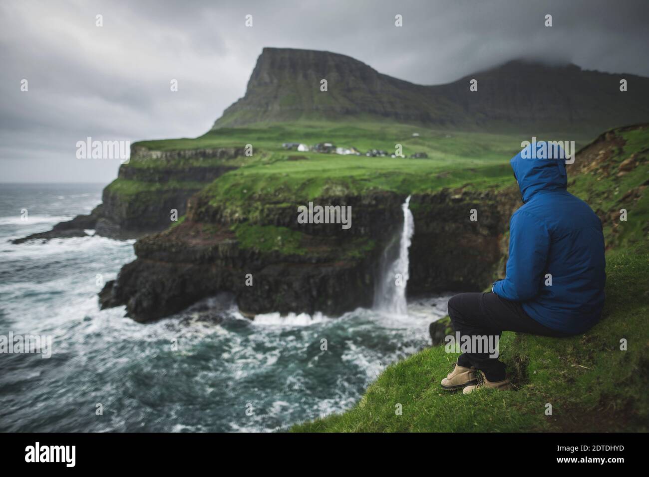 Denmark, Faroe Islands, Gasadalur Village, MÂ·Lafossur Waterfall, Man sitting on edge of cliff and looking at Mulafossur Waterfall Stock Photo