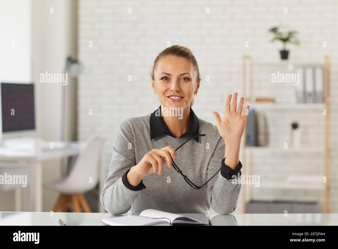 Happy teacher, student or businesswoman sitting at desk and waving hand at camera Stock Photo