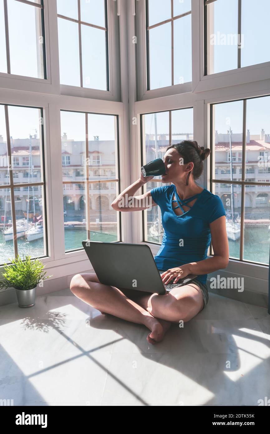 Young woman drinking coffee, working with laptop at the window at home over the marina. Freelance lifestyle and work at home concept. Stock Photo