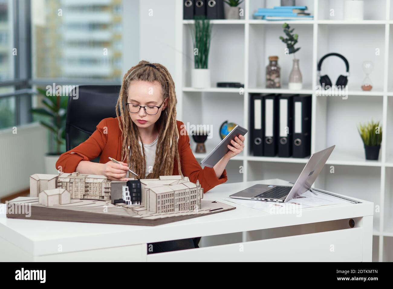 Female architect with dreadlocks makes measurements from mock-up of new buildings in architectural bureau. Stock Photo