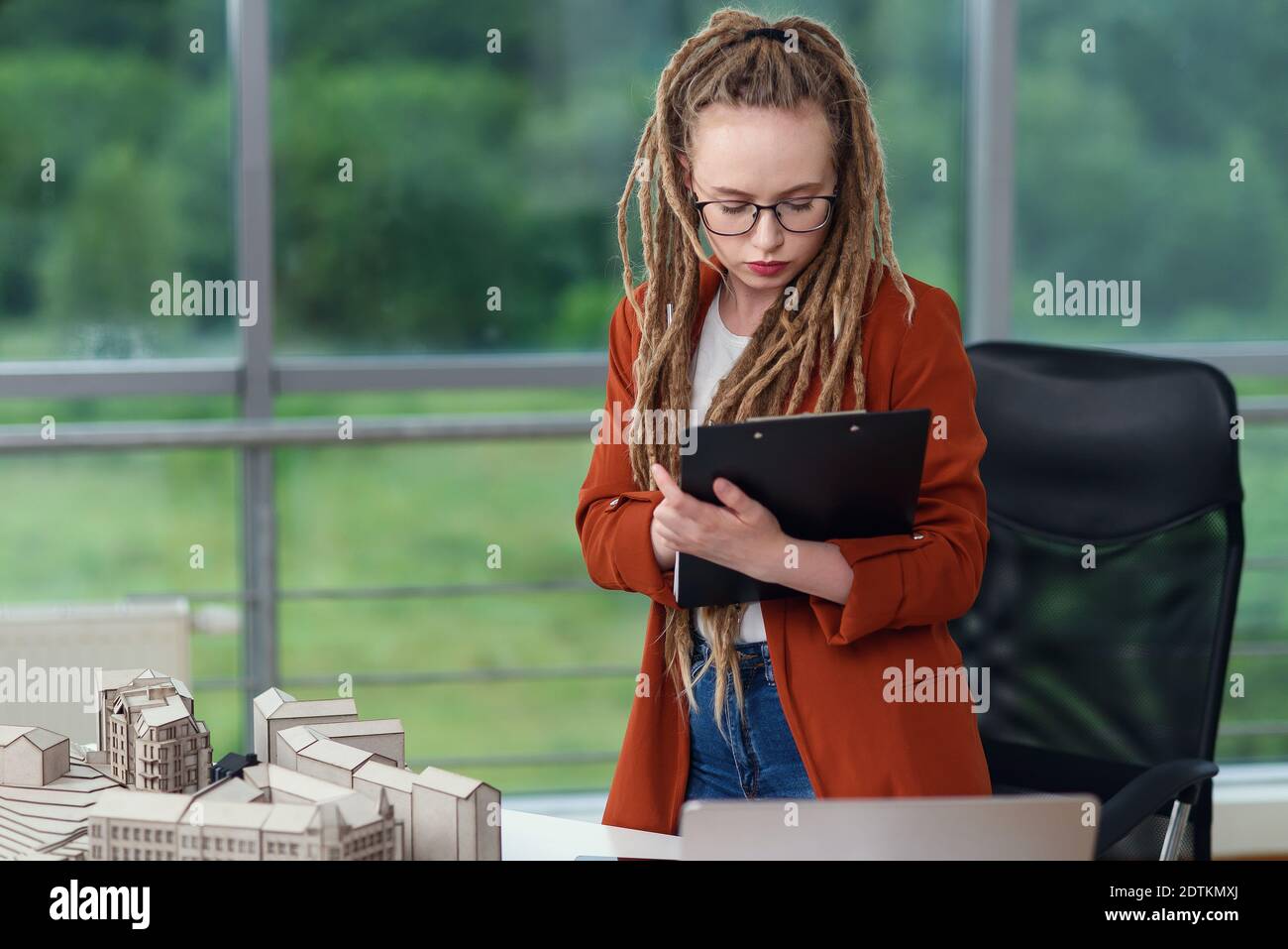 Adorable female engineer stands near constructed model and doing notes into the technical documents. Stock Photo