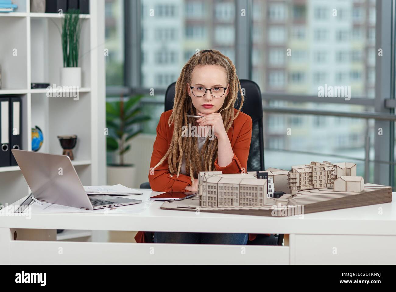 Stylish female architect in glasses with dreadlocks sits at her workplace with mock-up of future building. Stock Photo