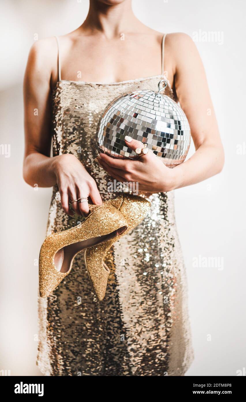 Young woman figure in festive glittering cocktail golden mini dress holding discoball and gold high heels shoes in hands over white wall. New Year holiday party concept Stock Photo