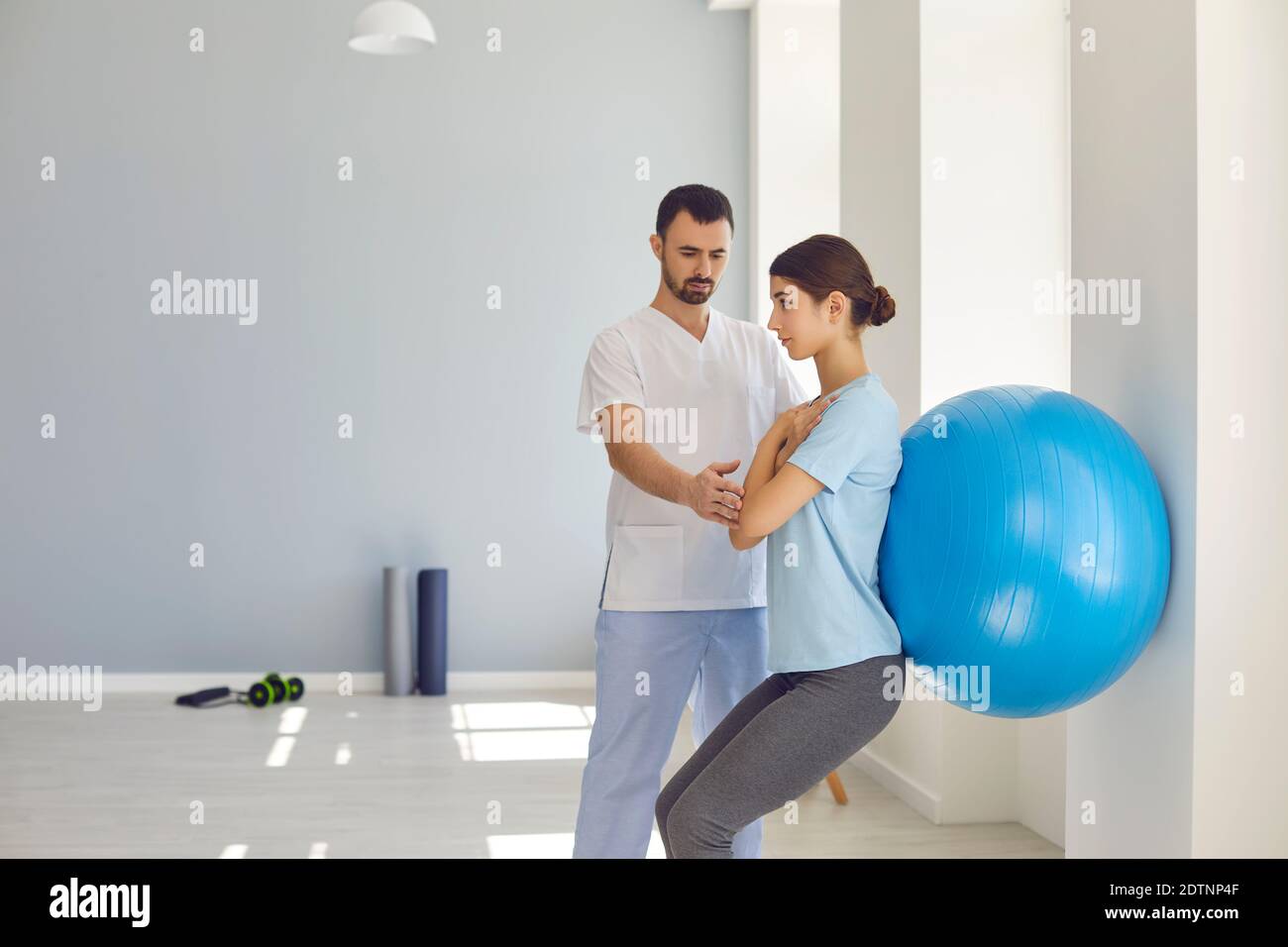 Physiotherapist helping young woman do wall squats with fit ball in modern clinic Stock Photo
