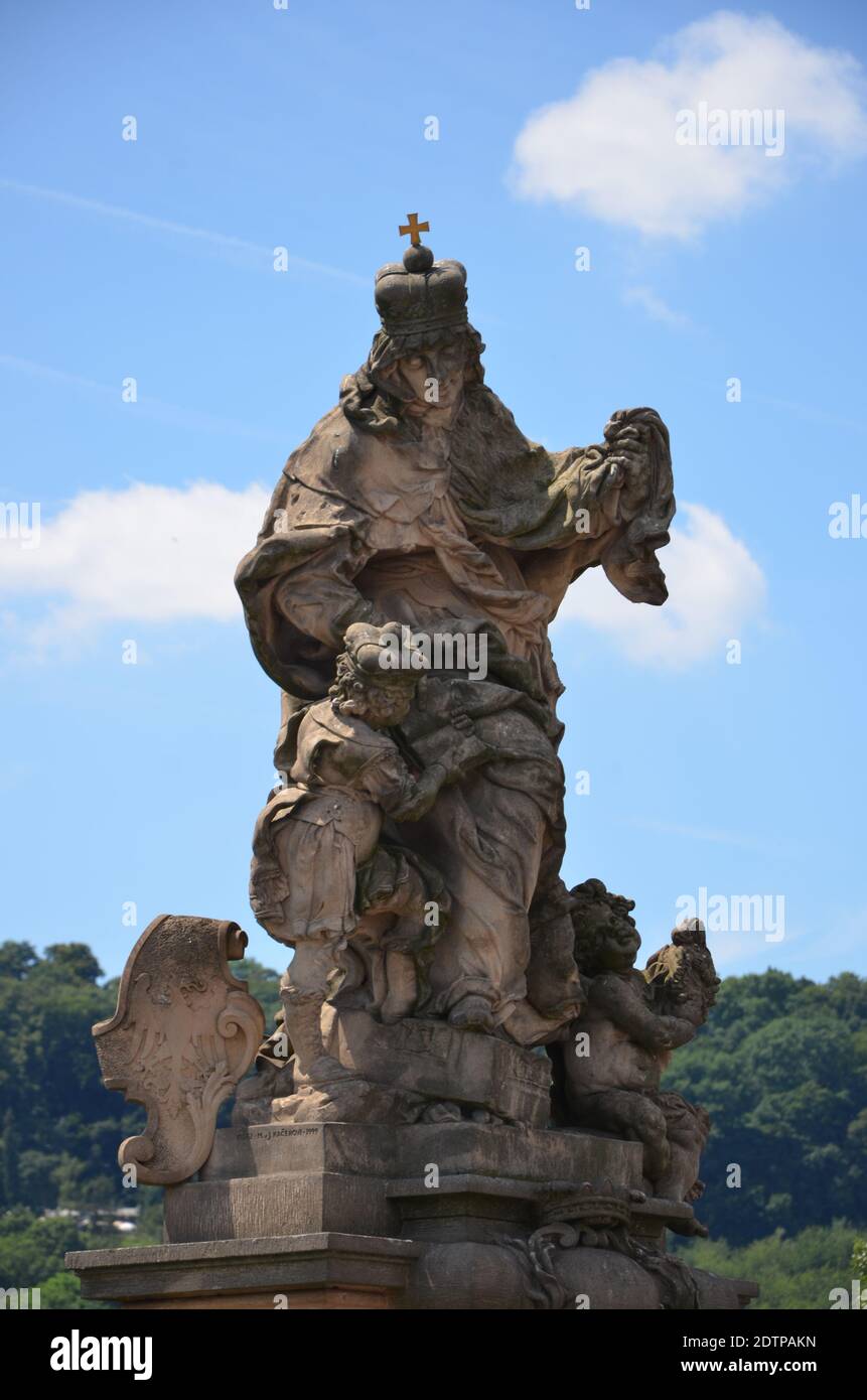 A historical statue decorated with religious figures and a meeting of green trees with a blue sky behind. Stock Photo