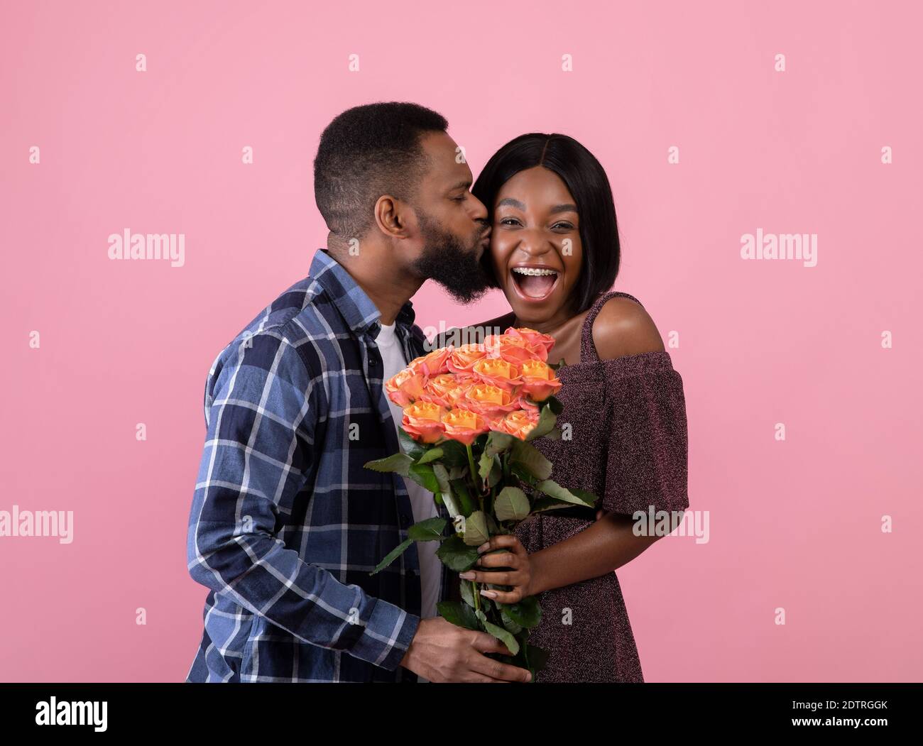 Affectionate African American guy kissing his happy girlfriend and giving her bouquet of roses on pink studio background Stock Photo