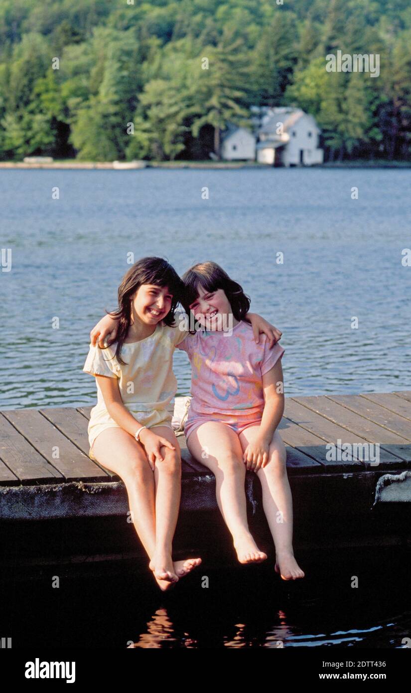 Two young girls on vacation with their parents sit on a wooden dock at a resort area on the Finger Lakes in the Adirondacks in upstate New York Stock Photo