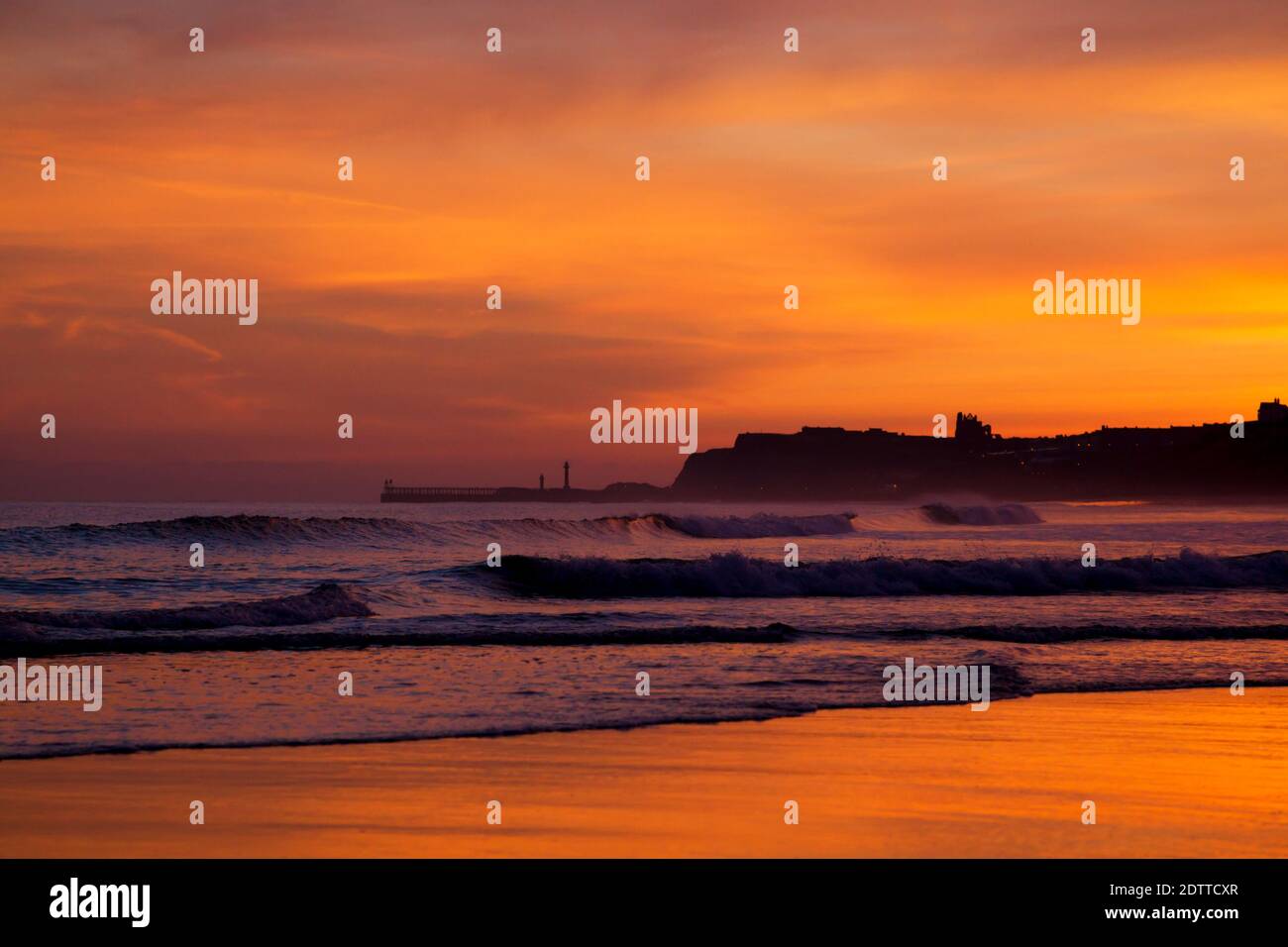 Daybreak view looking towards Whitby showing pier and abbey in silhouette against a colourful sky reflected on wet sand.  Viewed from Sandsend. Stock Photo