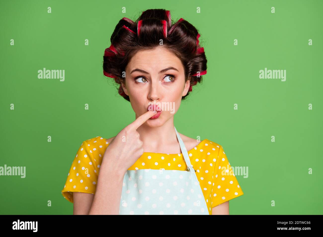 Photo of cute lovely young girl roller hairdo lips lick finger tasting cream look side thinking comparing with her previous try wear dotted apron Stock Photo