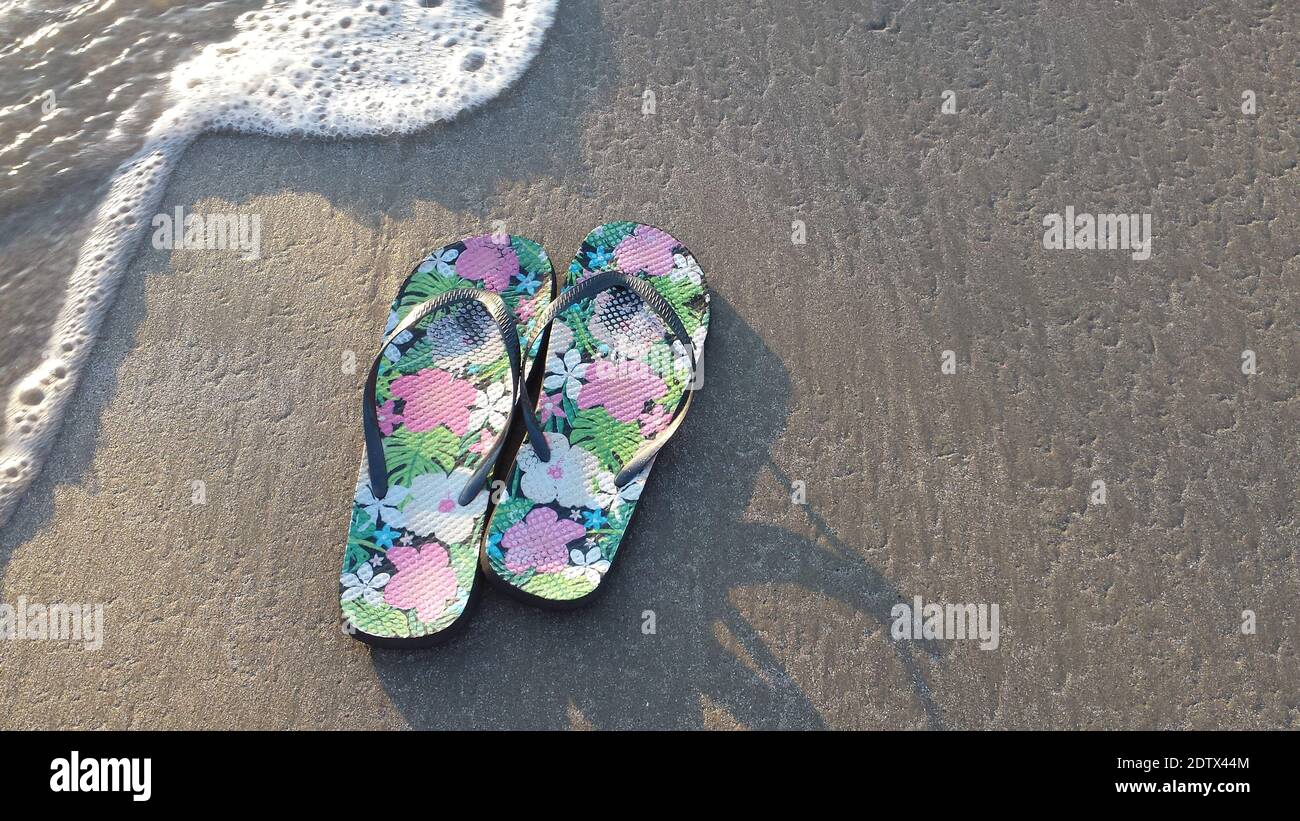 A pair of flowered flip-flops in the sand at the edge of the incoming tide, concept for summer, vacation, beach fun, tourism Stock Photo