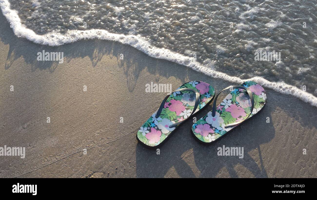 A pair of flowered flip-flops in the sand at the edge of the incoming tide, concept for summer, vacation, beach fun, tourism Stock Photo