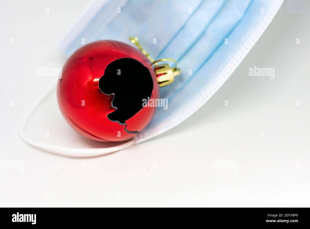 A broken decorative red Christmas tree bauble on a protective surgical mask on a white background. Christmas 2020 with the danger of the Covid-19 coro Stock Photo