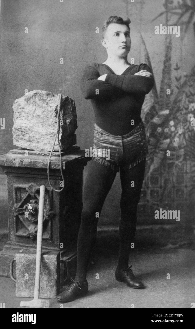 In this early-1890s photo, this fellow was a professional strongman who made his living doing feats of strength in circuses, sideshows, etc. The heavy block of stone has a rope attached, for lifting in various ways. At lower left is a flat, rectangular stone and a sledgehammer. One act involved putting the big stone on the strongman's chest, while he was lying down, and letting an audience member hit the stone with the sledgehammer.  There is a chance that this was Arthur Dandurand, a French-Canadian strongman.  To see my other circus-related images, Search:  Prestor  vintage  circus Stock Photo