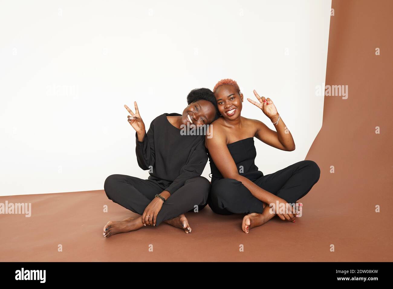 Portrait of stylish, energetic dark-skinned girls sitting on floor in light studio and posing with bright smiles Stock Photo