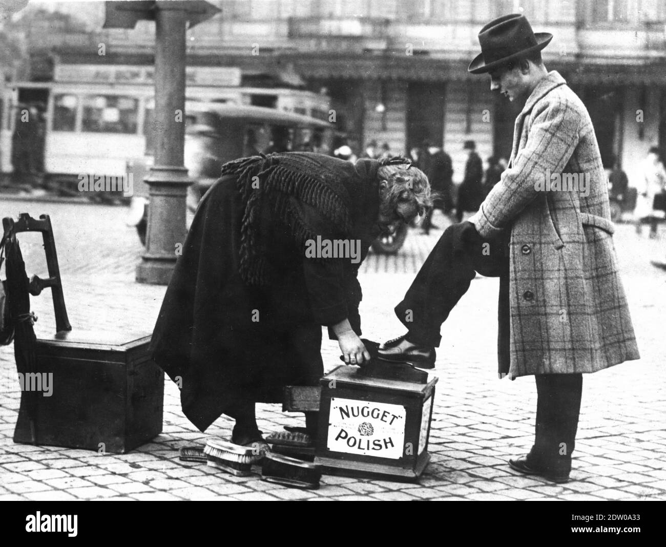 In a busy city street of Brussels, Belgium in the 1920s, a grey-haired old woman is bending over to shine the leather shoes of a young man.  She, hatless, is dressed in a black winter coat covered by a black shawl. He is dressed in a fine wool overcoat, homberg hat and black trousers. The woman has several brushes and a chair behind her. The man's foot is on a box clearly marked 'Nugget Polish'  The city street is quite busy, with public transit and people walking and waiting nearby.   To see my other interesting images of women, search:  prestor  vintage  woman Stock Photo