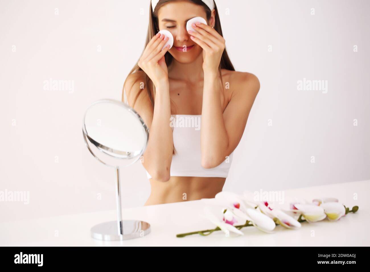 Young brunette girl takes care of her skin, standing in front of a mirror, enjoying beauty treatments for herself, smiling tenderly. Stock Photo