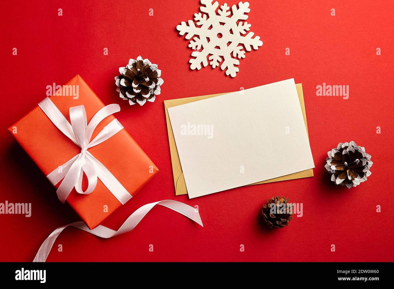 Christmas greeting card mockup. Gift box, pine cones and snowflake decorations on red paper background, top view, copy space Stock Photo