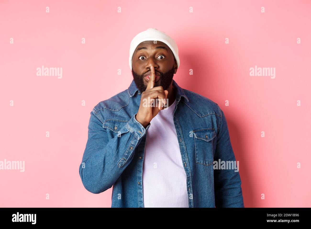 young black guy in beanie and denim shirt prepare hushing softly looking at camera, keep quiet, pink background Stock Photo - Alamy