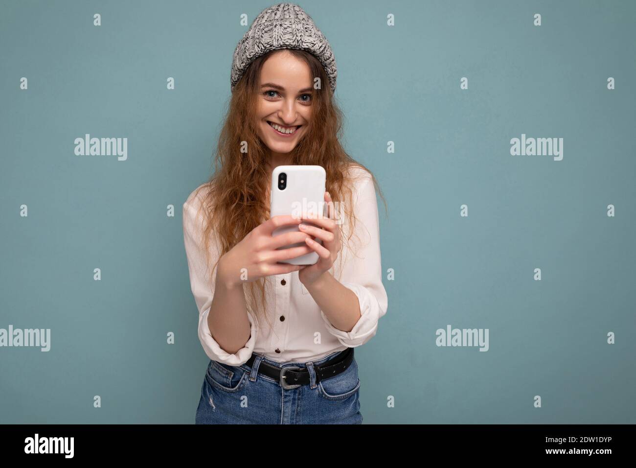 photo of attractive positive young blonde curly woman wearing casual white shirt and grey hat isolated over blue background wall holding phone and Stock Photo
