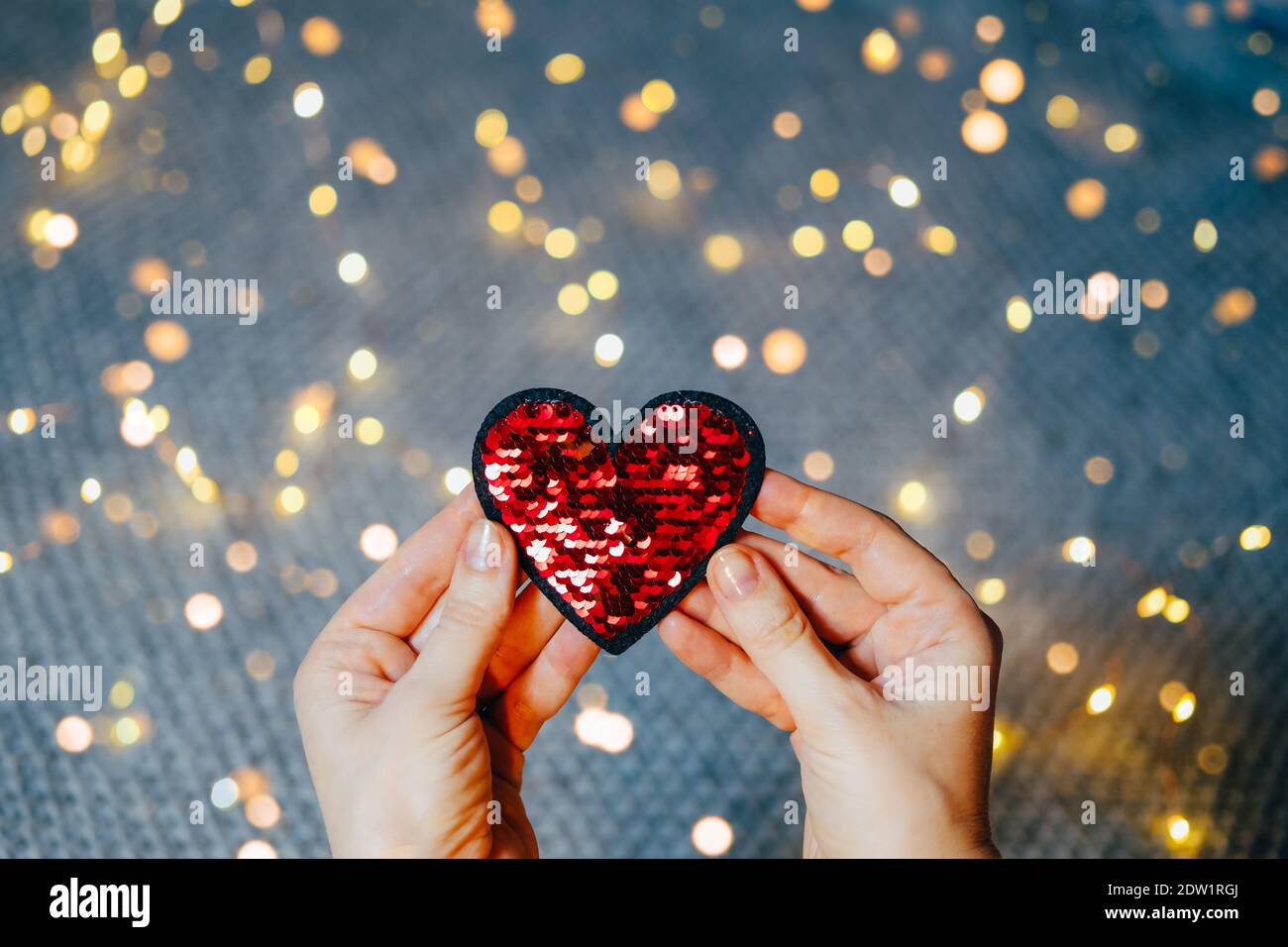 Red sequins heart in woman&rsquo;s hands on gray background with bokeh lights