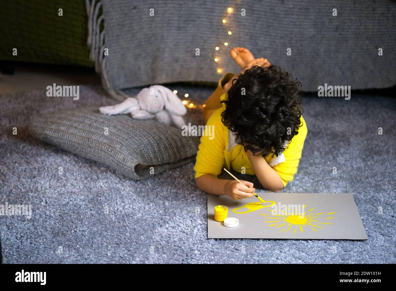 Little curly boy drawing yellow sun in gray interior at home. Stock Photo